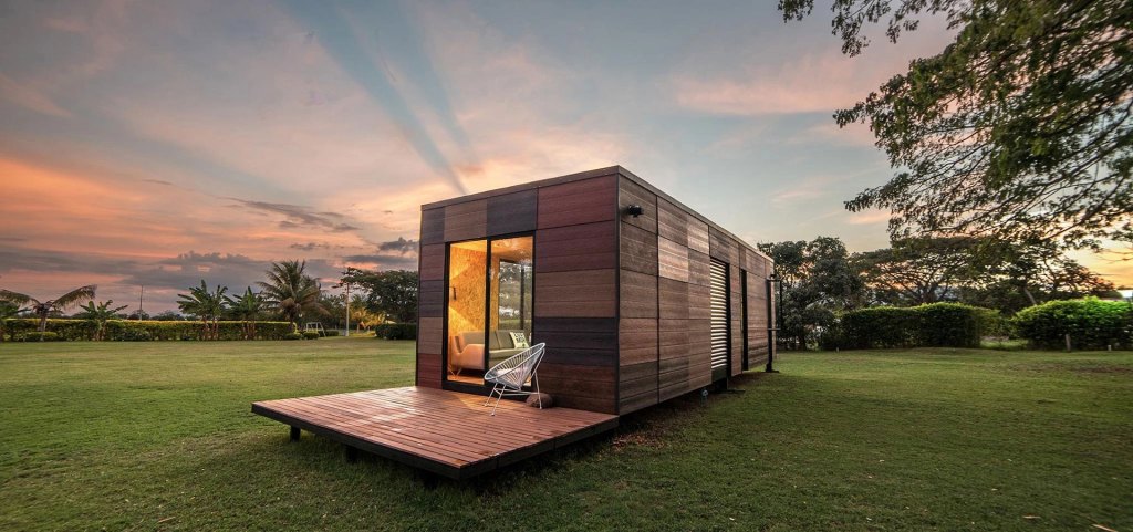 A small house surrounded by a grassy field under a blue sky with clouds, and trees in the background during sunset.