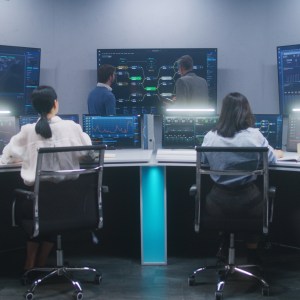 Female software engineer sits at computer with displayed blockchain network and surveillance maps. Technical support specialists work in monitoring room. Multiple big screens on the wall. Dolly shot.