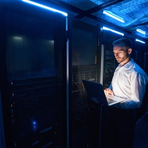 Caucasian man in data center standing near server racks with network equipment and using laptop